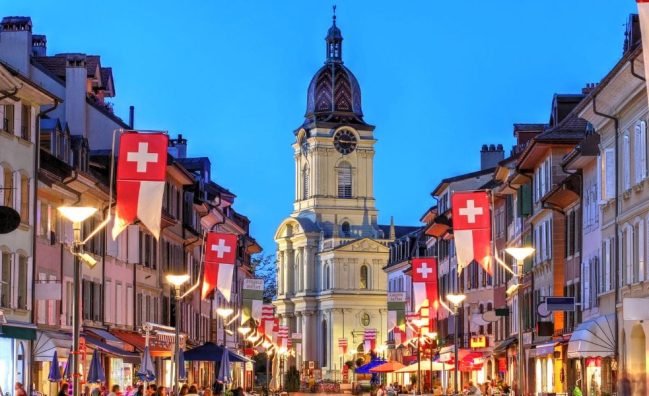 A city street in Morges with a clock tower in the background. Swiss flags flex banner hanging outside of restaurants walls. People are sitting on benches of street restaurants in Morges, Switzerland.