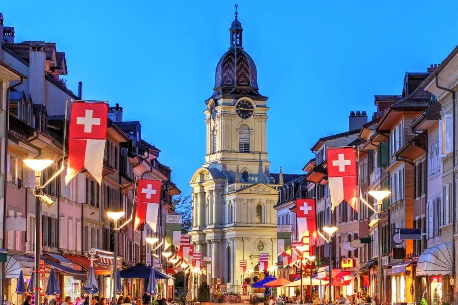 A city street in Morges with a clock tower in the background. Swiss flags flex banner hanging outside of restaurants walls. People are sitting on benches of street restaurants in Morges, Switzerland.