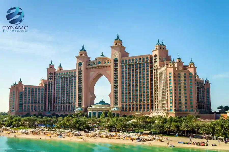Atlantis The Palm resort on Palm Jumeirah, Dubai, with grand archway, pink towers, teal roofs, sandy beach, palm trees, and turquoise waters under clear blue sky.