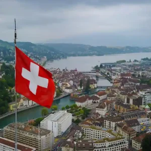 Travel poster featuring aerial view of Swiss city with large Swiss flag, river bridges, traditional European architecture, and mountain backdrop