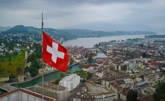 Travel poster featuring aerial view of Swiss city with large Swiss flag, river bridges, traditional European architecture, and mountain backdrop