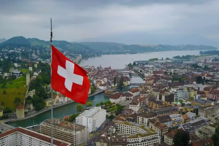 Travel poster featuring aerial view of Swiss city with large Swiss flag, river bridges, traditional European architecture, and mountain backdrop