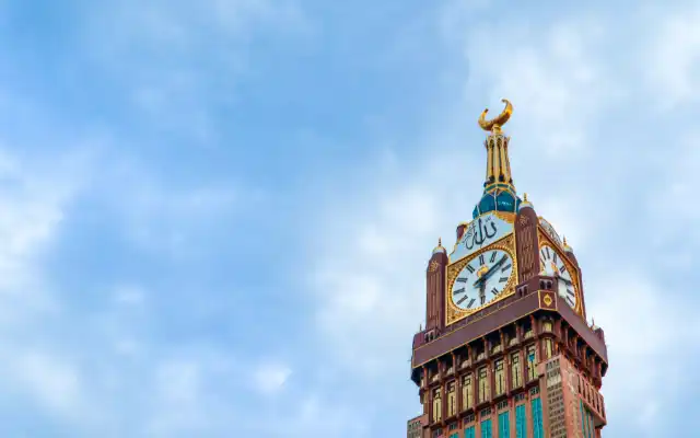 Top portion of the Abraj Al-Bait Clock Tower in Mecca featuring a large clock face with Arabic numerals, golden details, and crescent moon ornament.