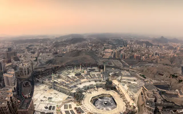 Aerial view of Masjid al-Haram in Mecca at sunset, showing the Kaaba at the center, surrounded by tall hotels and mountainous terrain