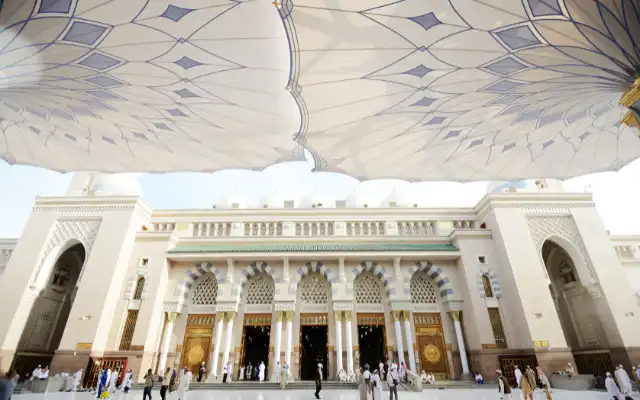 Exterior courtyard of Al-Masjid an-Nabawi in Medina with ornate doors, arches, and large retractable umbrella canopies providing shade for worshippers
