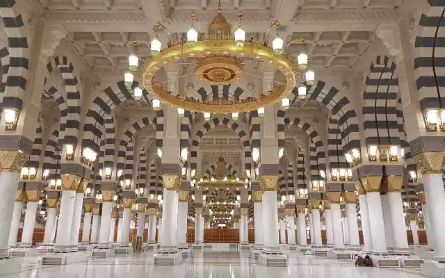 Interior of Al-Masjid an-Nabawi in Medina with tall marble columns, gold detailing, black-and-white patterned arches, and grand chandeliers - Umrah packages from Pakistan