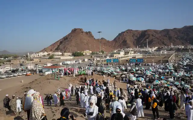 Large crowd of pilgrims in white garments gathered at Mina during Hajj, with rocky hills, tents, and vehicles visible in the desert background
